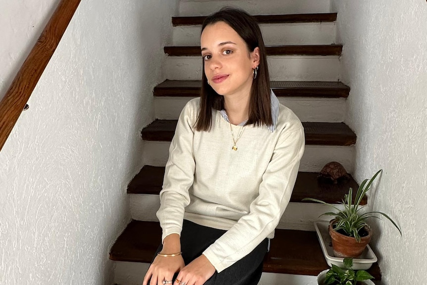 A young woman with straight dark brown shoulder-length hair sitting on stairs next to house plants.
