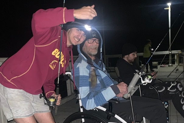 David Mobbs sits in a wheelchair while fishing on a bridge at night next to a man standing in a red jumper.