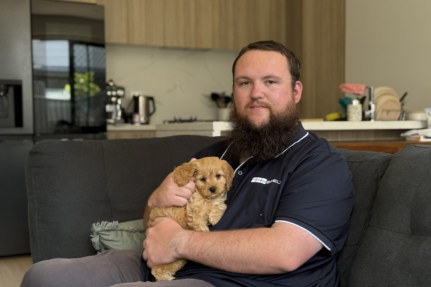 man with beard in polo shirt holds small puppy on a couch in front of a kitchen