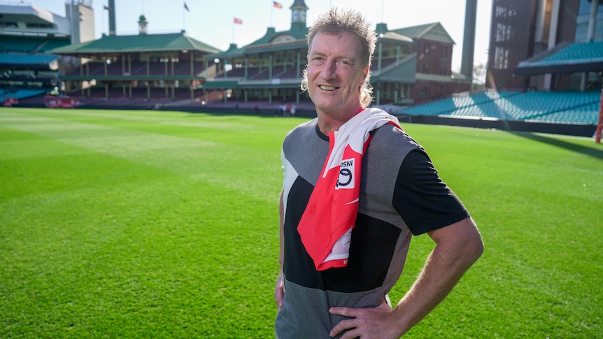 A former AFL footballer stands in the middle of the SCG smiling at the camera and holding a guernsey.
