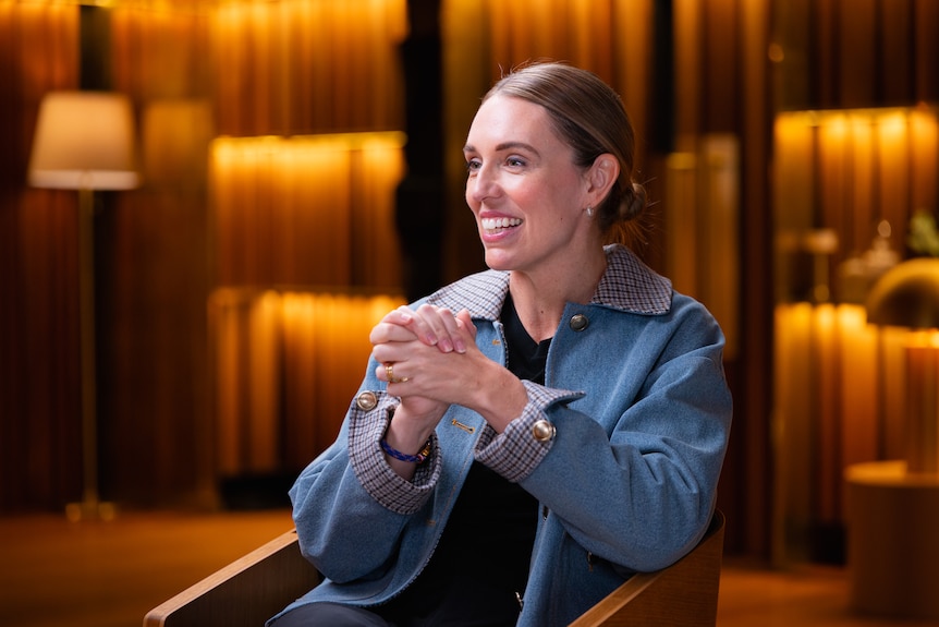 Jacinda Ardern, in a denim jacket with hair pulled back in low bun, sits in a warmly lit room talking with her hands clasped