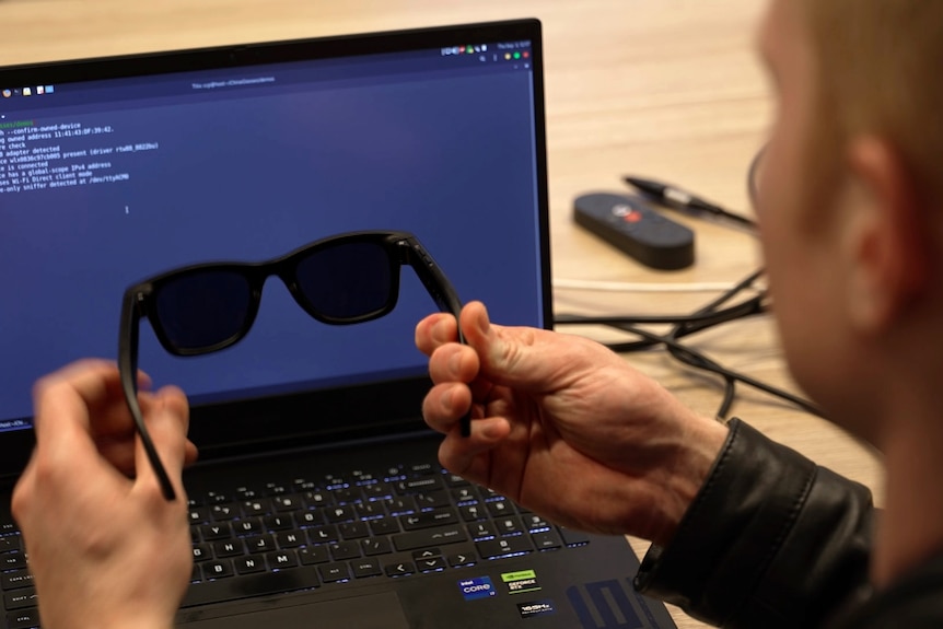 A man seen from behind holds a pair of black sunglasses in front of a computer laptop screen