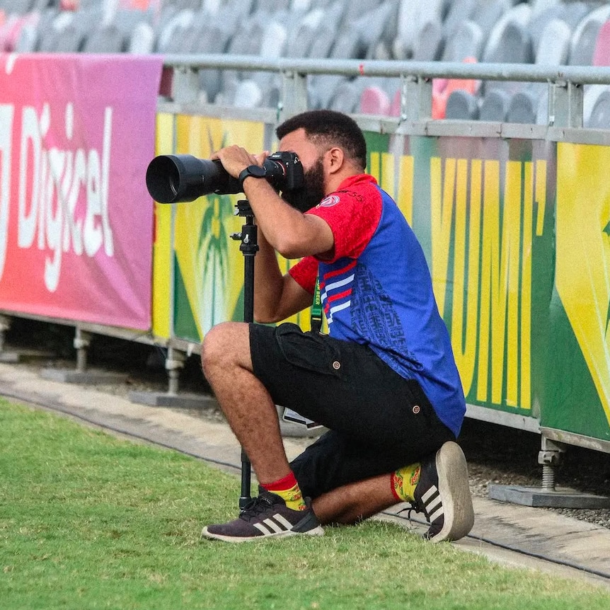 A PNG man wearing a blue and red top and black shorts kneels on a field on one knee behind a camera mounted on a mono-pod