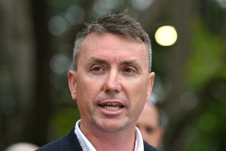 a close-up image of a white man speaking with green shrubbery in the background