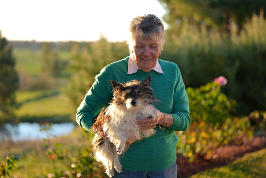 An older woman with grey hair cuddles her small dog while standing in her pretty garden.