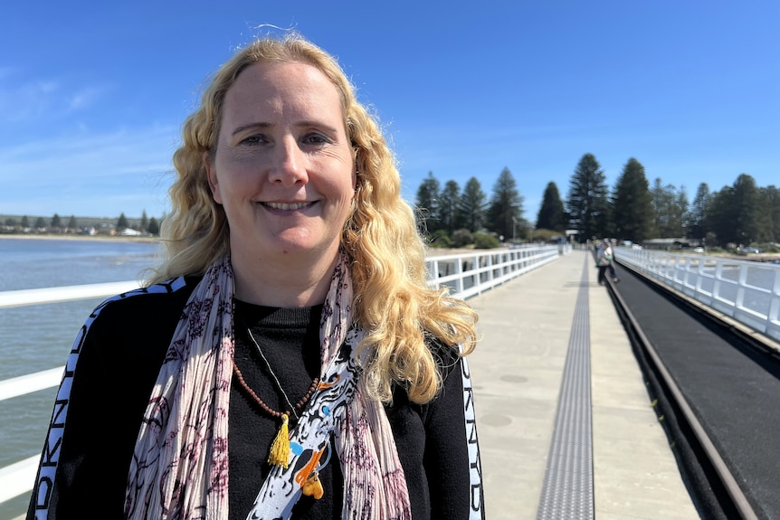 A woman with long blonde hair standing on a bridge over the sea with Norfolk pines on the land behind her