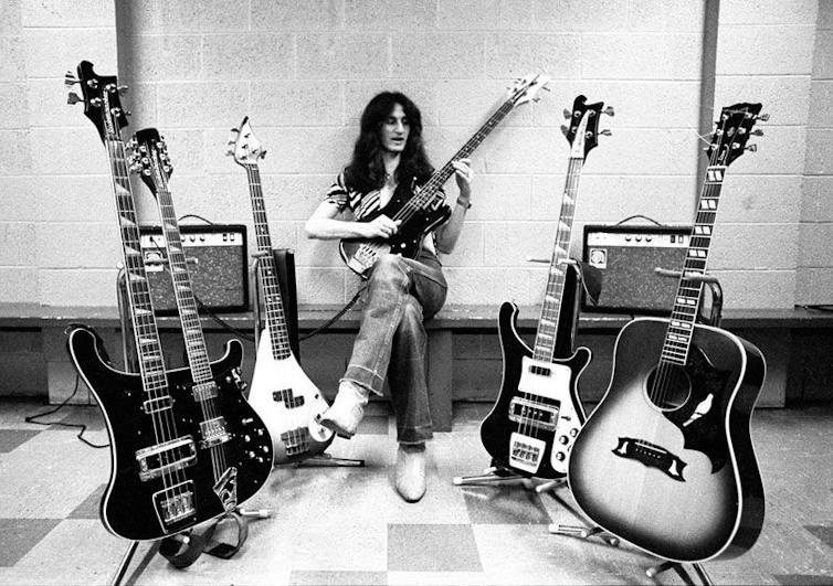 A man in cowboy boots with long hair sits amid guitars.