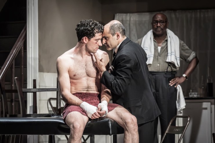 A boxing manager in a suit talks to a shirtless boxer with bandaged hands, while another man watches in the background.