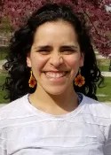 A person with long dark curly hair smiles outdoors, wearing a white shirt and colorful beaded flower earrings, with pink blossoms and green grass in the background.