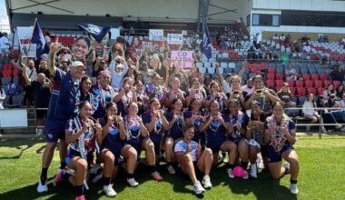 A large group of young girls in rugby league uniform pose and smile for the camera in front of a grandstand