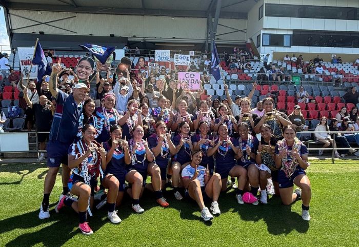 A large group of young girls in rugby league uniform pose and smile for the camera in front of a grandstand