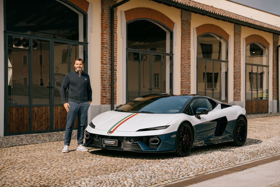 A man stands beside a white and blue Lamborghini Temerario Tricolore sports car parked outside a brick building.