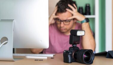 A person wearing glasses sits at a desk with their hands on their head, looking stressed, with a camera and flash in the foreground and a computer monitor to the left.