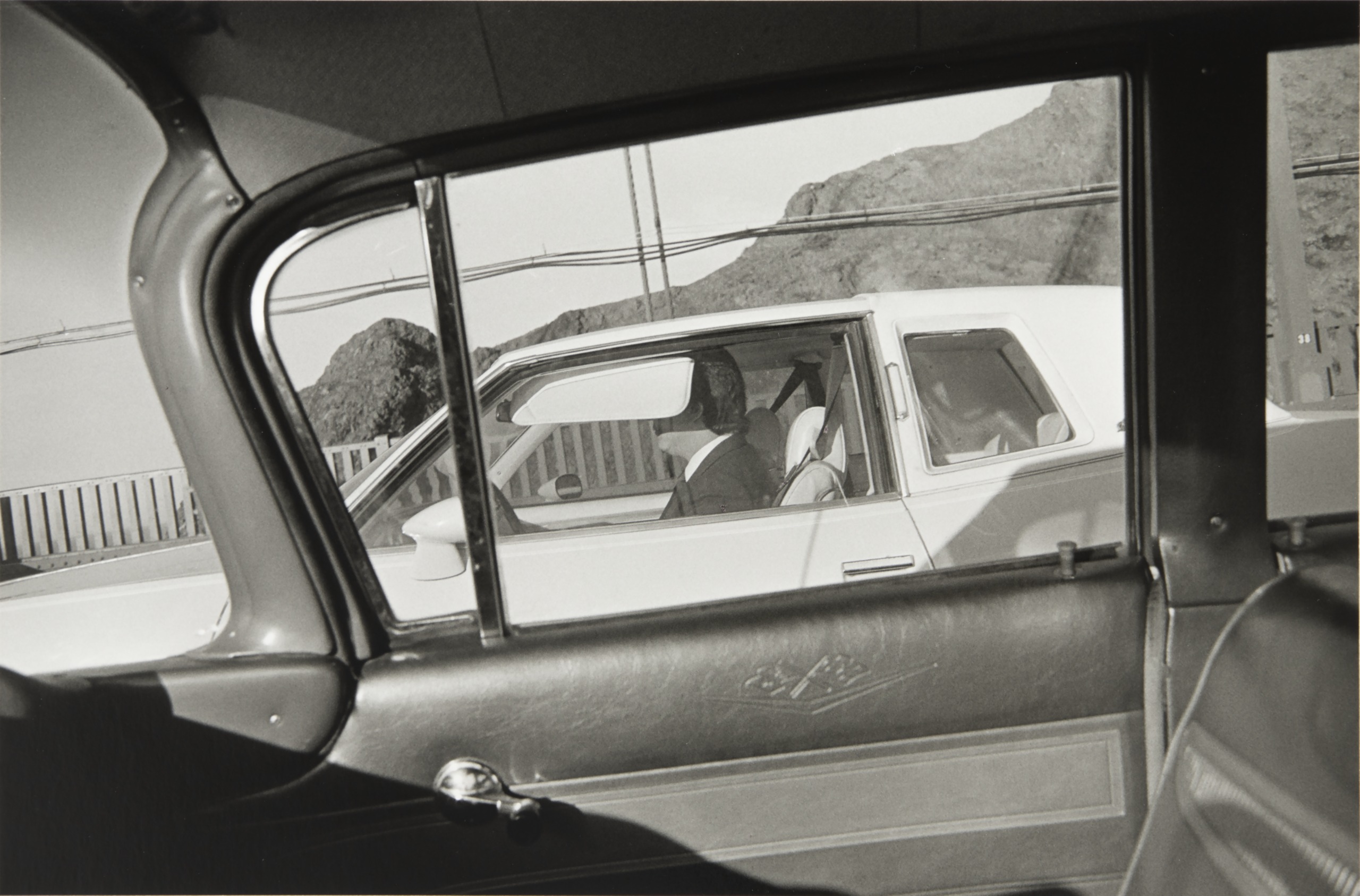 A black and white photograph taken from inside one car, framing a driver seen through the window of a neighbouring white car on a mountain road.