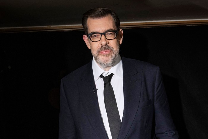 Richard Osman smiling ahead wearing a black suit and tie.