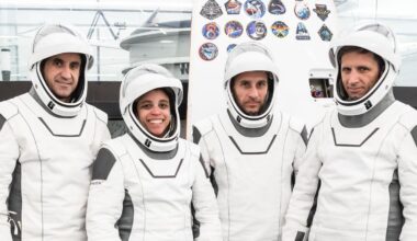 three male and one female astronauts in white flight suits stand in a big, white-walled room