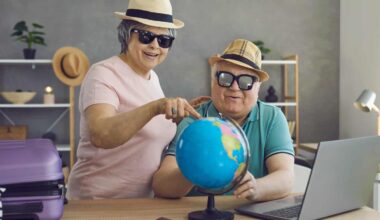 A smiling older woman in a pink shirt, straw hat, and sunglasses points at a blue globe while an older man in a green polo shirt, straw hat, and sunglasses holds the globe. A purple suitcase and passports are visible on a wooden desk with a laptop in a home setting.