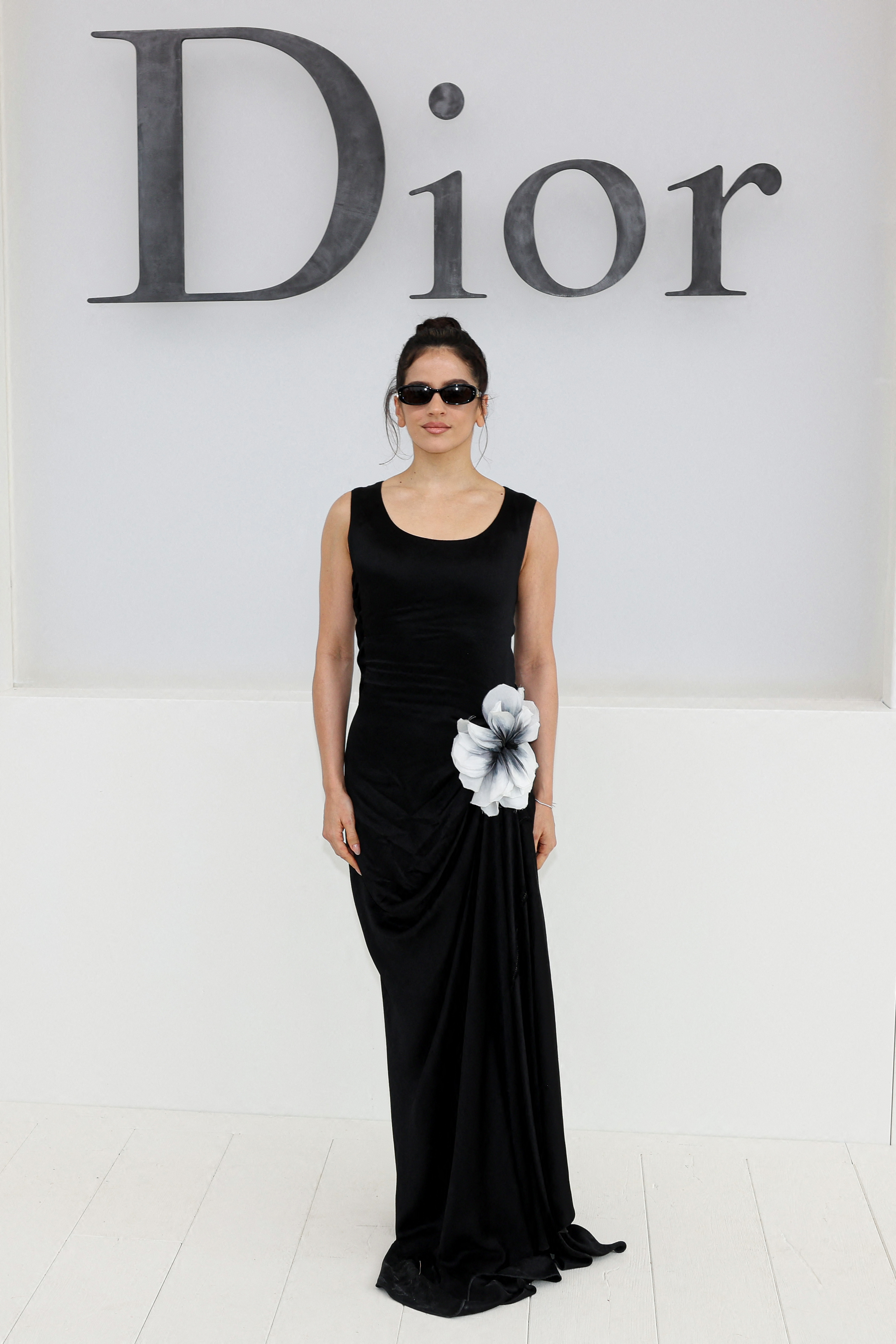 Rosalía poses for a photocall in a black dress with a large black and white flower embellishment, against a white wall with the Dior logo.
