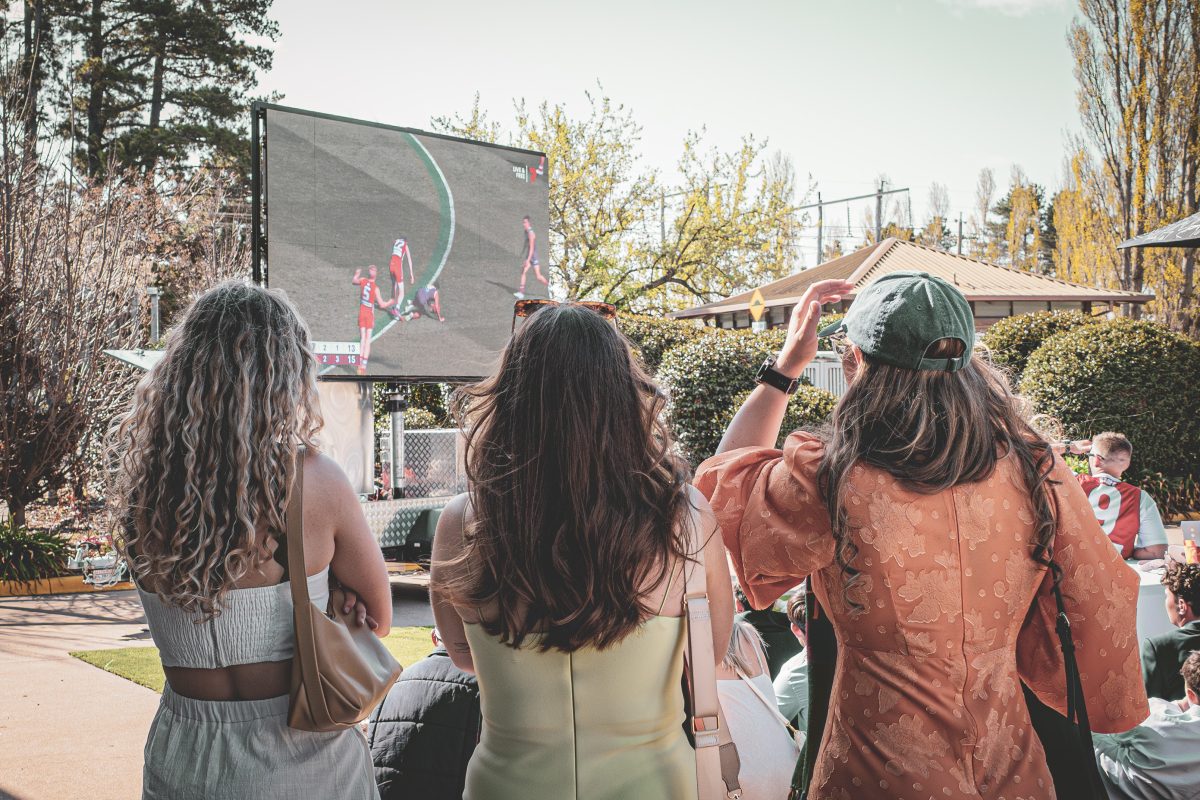 people standing watching a large screen playing AFL Grand Final outside