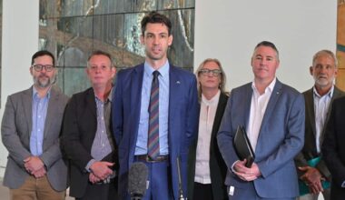 A tall man in a blue suit and striped tie stands at a microphone speaking to reporters, flanked by five other people in professional attire.