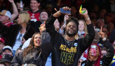 In a crowded stadium stand, a smiling man with dreadlocks wearing a black jersey with "MELBOURNE NFL" in yellow lettering raises both arms in excitement.