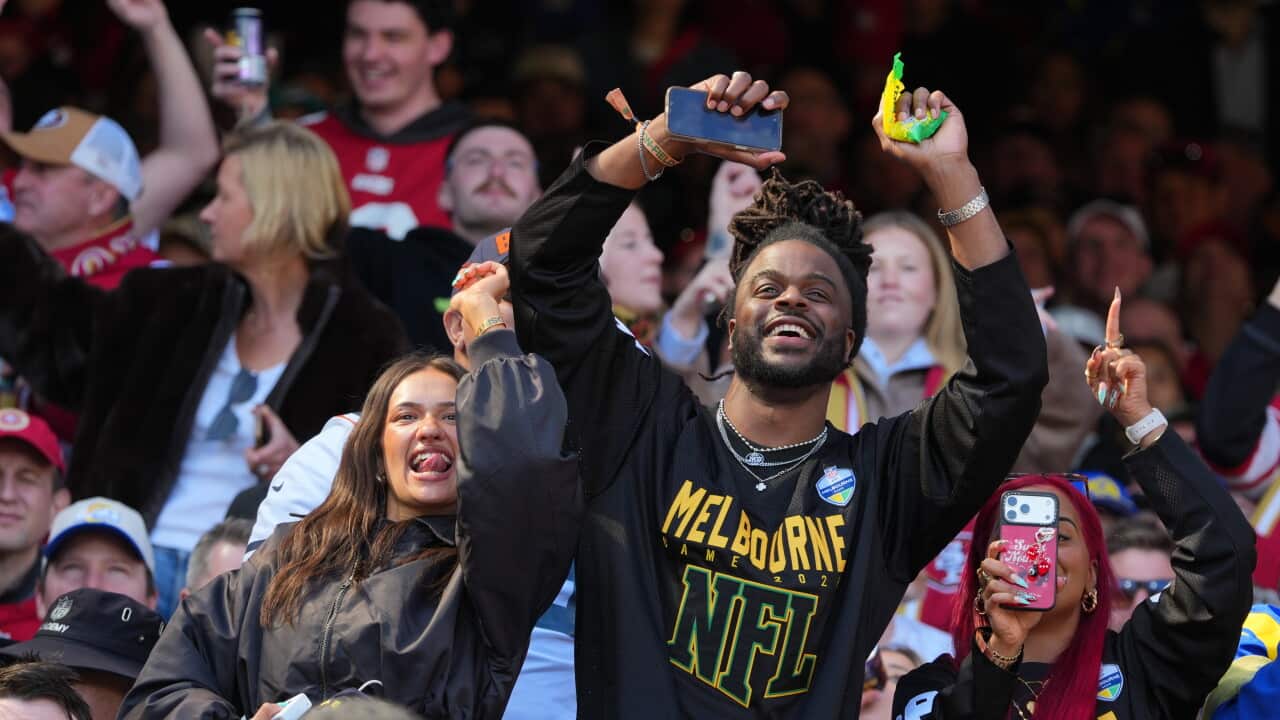In a crowded stadium stand, a smiling man with dreadlocks wearing a black jersey with "MELBOURNE NFL" in yellow lettering raises both arms in excitement.