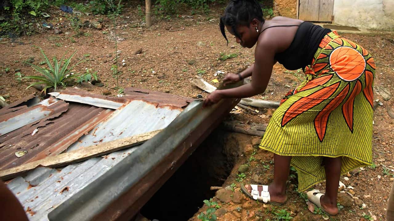 A woman in a black tank top and a patterned orange and yellow skirt lifts a large sheet of corrugated metal covering a hole in the ground.