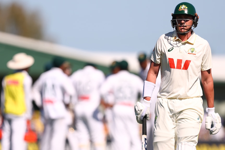 A cricket batter in white walks off the field carrying bat, with opponents celebrating behind him