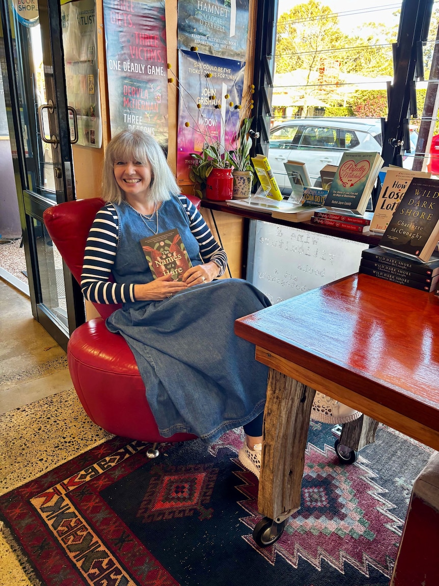 Bernice Barry, an older woman, sits in a bookshop, smiling, holding her book, featuring hares on the cover, to her chest.