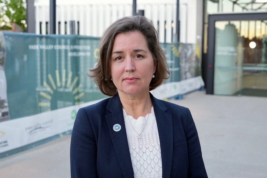 A woman with short dark hair wearing a blue blazer stands outside a building.
