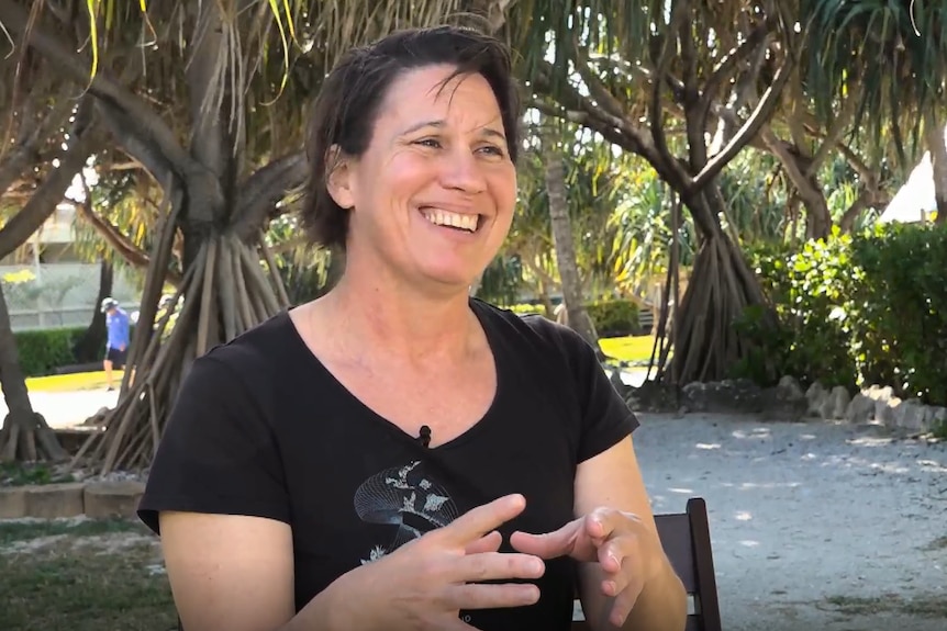 Christine, a smiling woman with short brown hair, sits on a chair in front of pandanus trees.