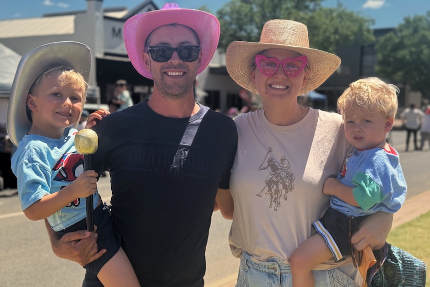 A couple wearing broad-brimmed hats and sunglasses stand near a street on a sunny day, both holding their young children.