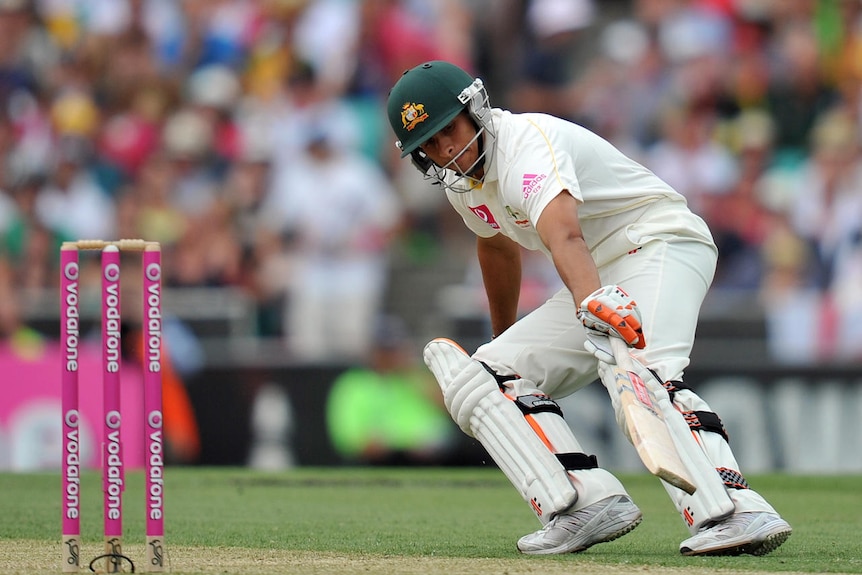 Usman Khawaja wearing cricket whites and a helmet leans down as he runs on the pitch. A crowd is in background.