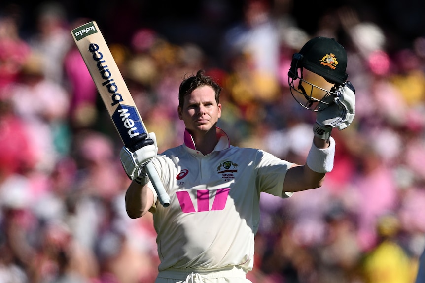 A cricket batter holds up his helmet and bat in celebration of a century