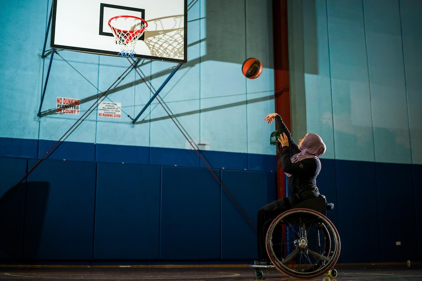 Reema El Qassem, wearing a hijab, sits in her wheelchair and shoots a basketball towards a hoop.