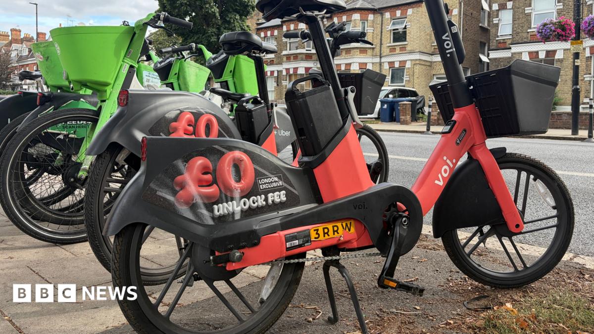 Image of a red Voi e-bike and green Lime bike parked on the side of a pavement. In the background there are rows of houses.