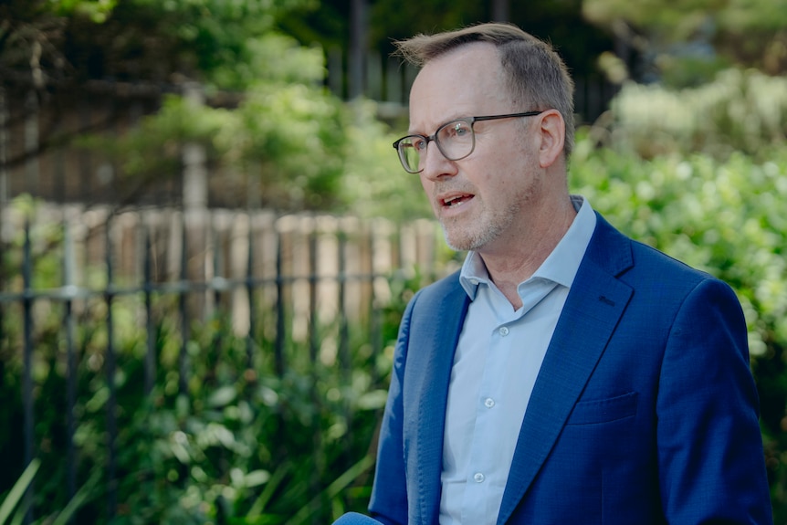 A side shot of Mr Shoebridge wearing a blazer and glasses talking into microphones outside.