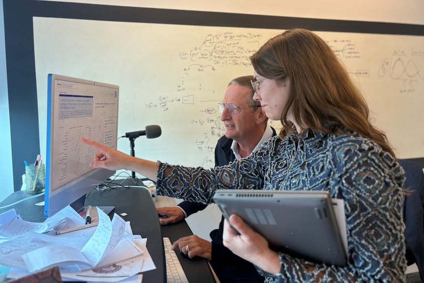 A woman and a man point to a computer screen covered in workings.