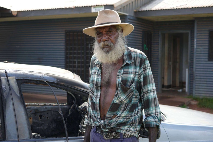 An older Indigenous man standing next to a burnt out car.