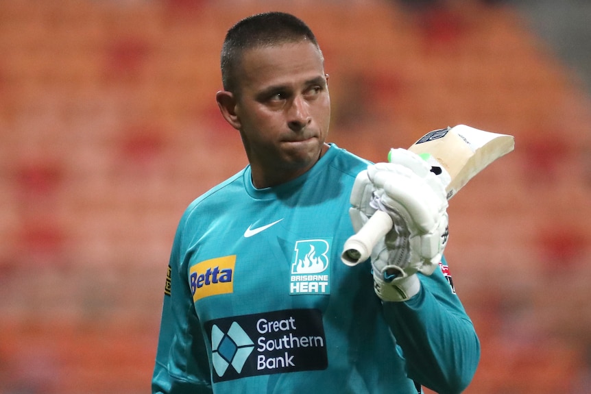 Usman Khawaja looks serious as he holds a cricket bat over his shoulder. He wears an aqua-coloured shirt.