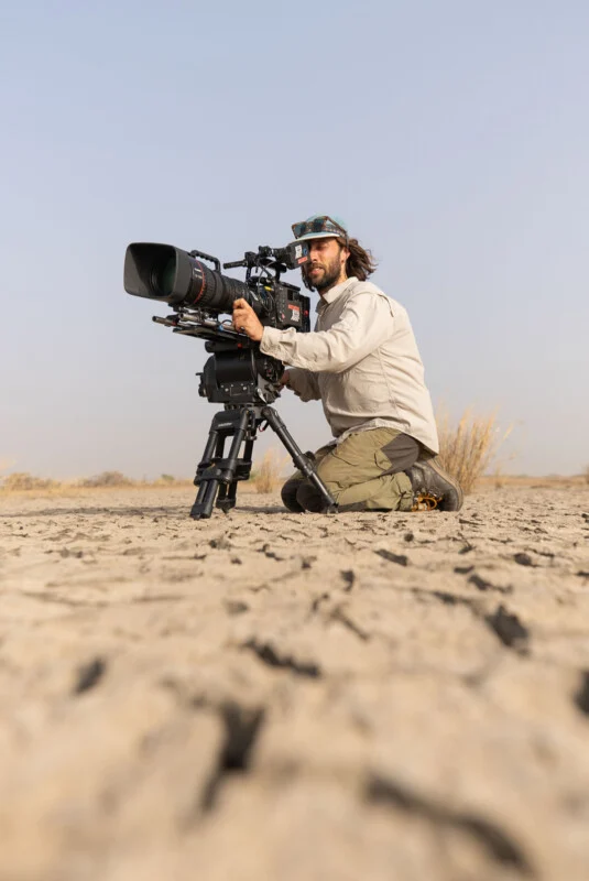 A man with long hair and a cap kneels on cracked, dry earth while operating a professional video camera mounted on a tripod.