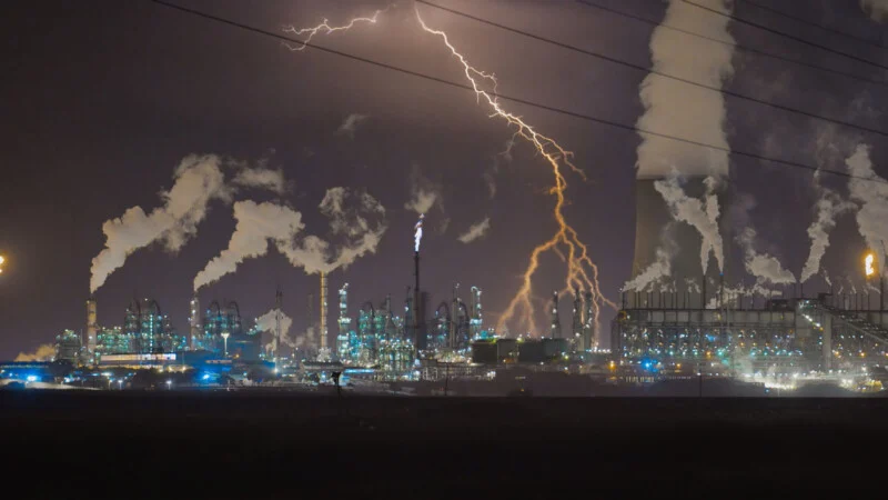 A lightning bolt strikes near an industrial plant at night, with smoke rising from multiple chimneys and cooling towers.
