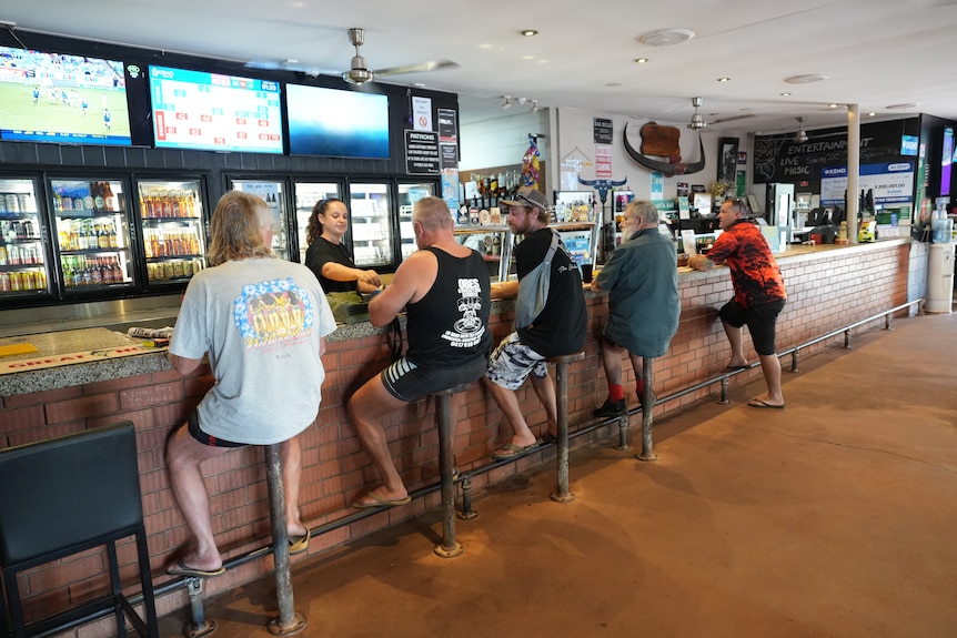 Men in shorts and thongs sit at a bar that is being tended by a woman in a dark shirt.
