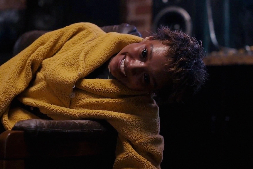 A woman of colour with short dark hair wears a yellow jacket and lens on a lounge arm; her eyes are dilated black.