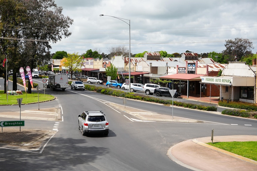 A road through a town, with shops on the right-hand side and a sign pointing left to "Broughton".