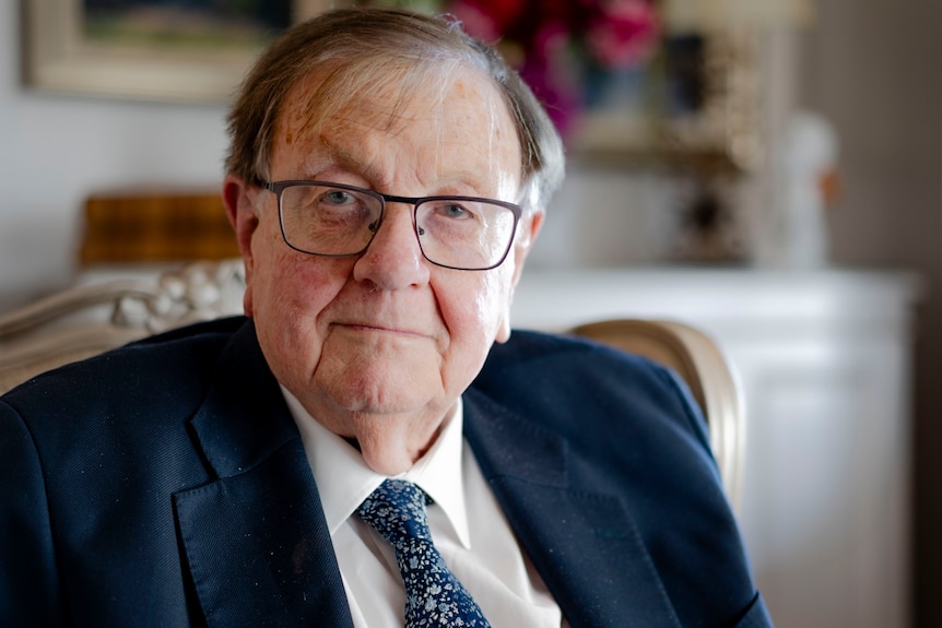 An older man in a suit with glasses, smiles sternly at a table.