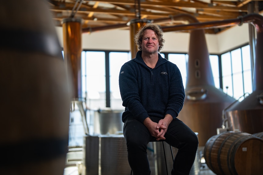 A man with curly hair sits surrounded by large copper distilling equipment