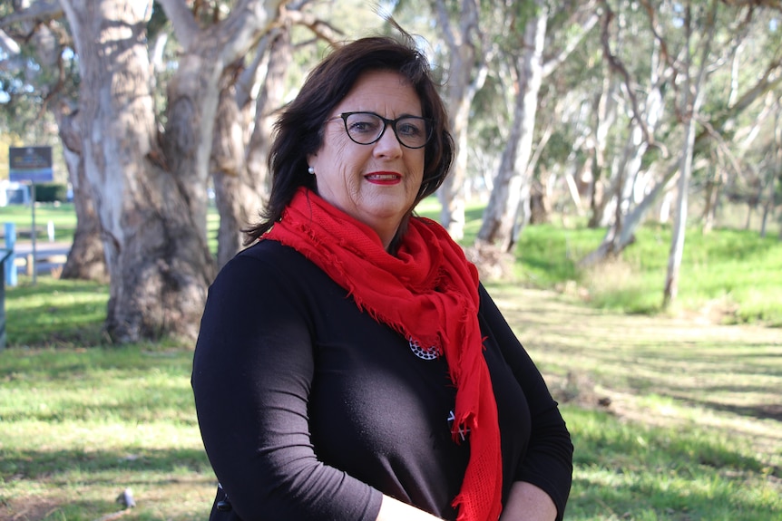 A woman in a red scarf with dark hair smiles at the camera, trees and grass behind her