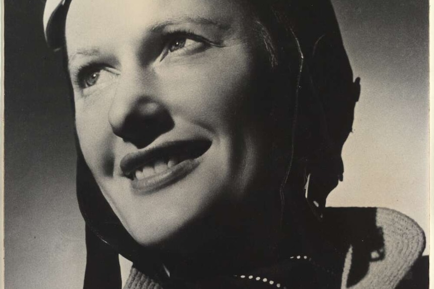 A black and white portrait of a woman in flying hat and goggles smiling looking up to the light.