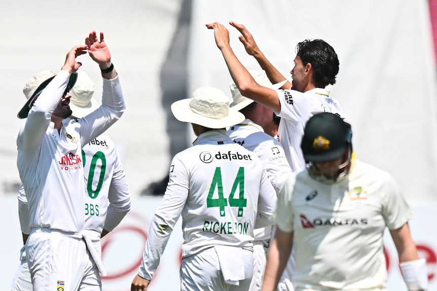 A cricketer in white walks off the field after being dismissed, players in white celebrating behind him